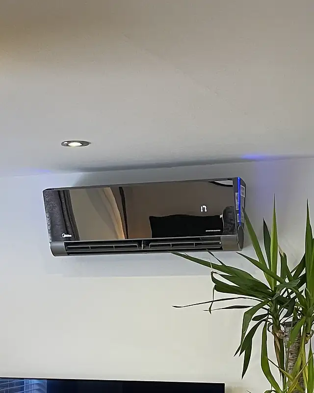 Mirror-finish wall-mounted air conditioner, London Mirror-finish black wall-mounted air conditioning unit with a softly lit display, mounted high on a living room wall beside a yucca plant